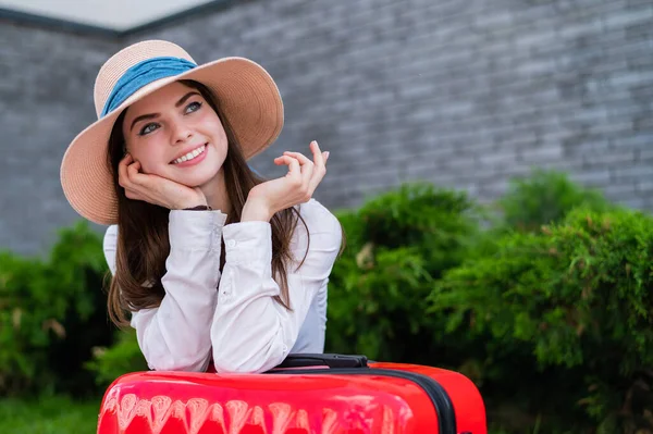 Portret van een mooie blanke vrouw in een hoed met een grote rode koffer buiten. Aantrekkelijk meisje leunde tegen bagage tijdens het wachten op een taxi. — Stockfoto