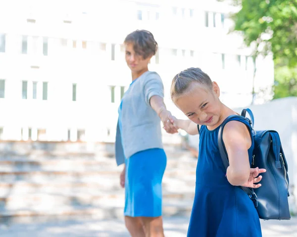 Uma menina infeliz com uma mochila puxa a mãe pela mão não quer ir à escola. Uma mulher leva uma filha que resiste à escola. O conflito de gerações. — Fotografia de Stock