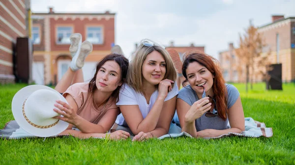 Tre belle giovani donne caucasiche si trovano sul prato in campagna. La bruna bionda e la rossa stanno riposando nel parco sull'erba verde. Gli amici hanno fatto un picnic all'aperto. — Foto Stock