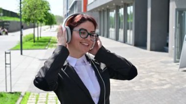 Takım elbiseli beyaz iş kadını kulaklıkla müzik dinliyor ve açık havada dans etmek için şarkı söylüyor.
