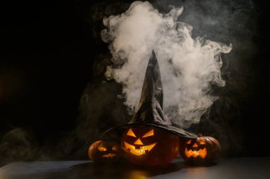 Sisin ortasında karanlıkta üç tane balkabağı feneri parlıyor. Cadılar Bayramı kabağı ve cadı şapkası..