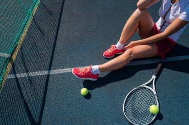 Spor etekli yüzü olmayan bir kız tenis kortunda oturuyor ve elinde bir roket tutuyor. Kadın bacaklarının üst görüntüsü.