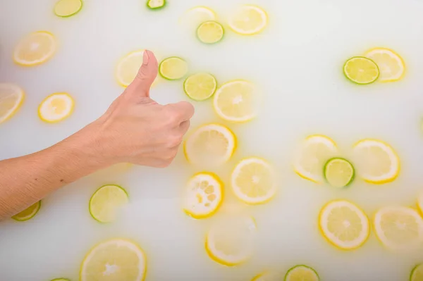 Une main de femme montre son pouce sur le fond d'un bain de lait aux agrumes. Femme gestes approbation tout en prenant un bain avec des citrons et des citrons verts. — Photo