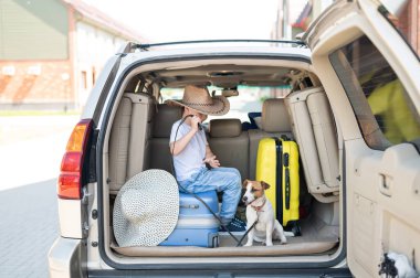 Kovboy şapkalı mutlu çocuk ve köpek yavrusu Jack Russell Terrier arabayla seyahat ediyor. Bir çocuk ve komik küçük bir köpek bagajda oturuyorlar ve yaz tatiline hazırlar. Bağımsız seyahat. En iyi arkadaşlar.