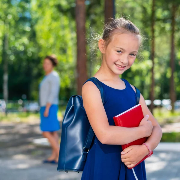 Porträtt av en flitig skolflicka med ryggsäck och en bok går i första klass. Mamma skickar en lycklig dotter till grundskolan. — Stockfoto