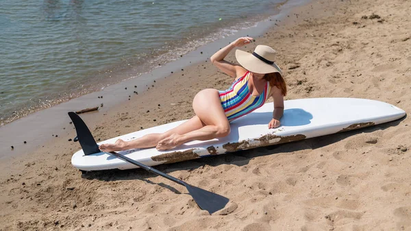 Egy nő fürdőruhában és kalapban pózol egy szörfdeszkán a parton.. — Stock Fotó