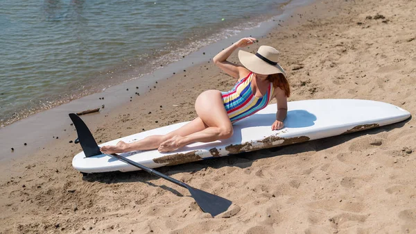 Eine Frau in Badeanzug und Hut posiert auf einem Surfbrett am Strand. — Stockfoto