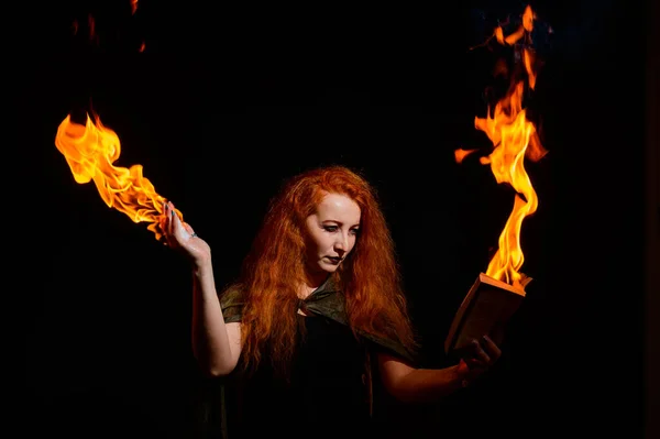 An ominous witch sets her spell book on fire with the power of thought. Red-haired woman conjures for Halloween. Flames on the hands.