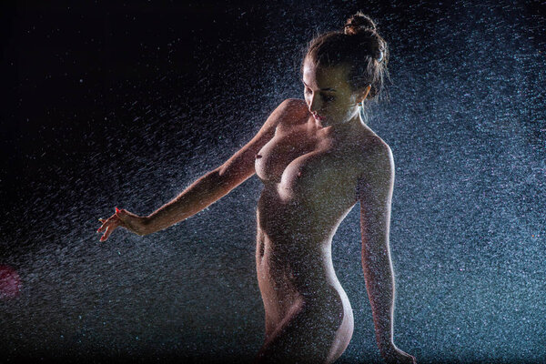 Прекрасная европейская обнаженная женщина стоит на лужайке под разбрызгивателем ночью. Вспышки освещают девушку без одежды в каплях лужайки с водой. 