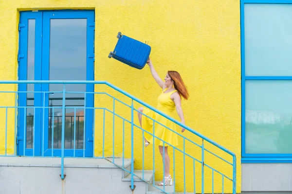 Feliz mujer pelirroja con un vestido amarillo sube con una maleta azul. —  Fotos de Stock