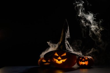 Sisin ortasında karanlıkta üç tane balkabağı feneri parlıyor. Cadılar Bayramı kabağı ve cadı şapkası..