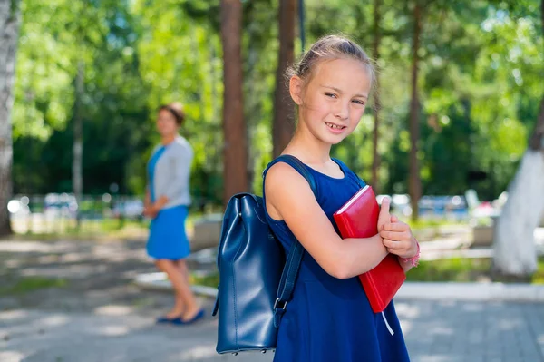 Portret sumiennej uczennicy z plecakiem i książką idzie do pierwszej klasy. Mama wysyła szczęśliwą córkę do szkoły podstawowej. — Zdjęcie stockowe