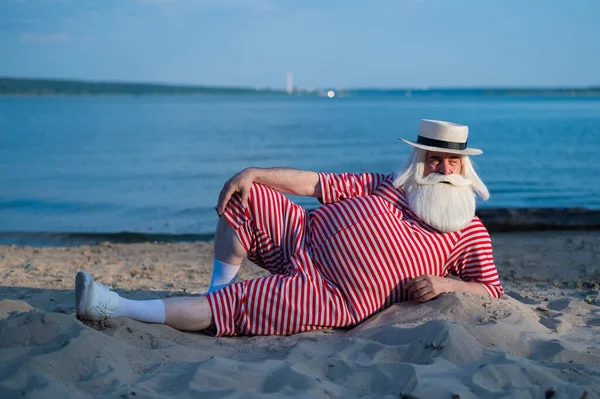 Un uomo anziano in costume da bagno retrò a righe prende il sole sulla spiaggia. Un vecchio uomo barbuto dai capelli grigi con un cappello giace sulla sabbia vicino al mare. — Foto Stock