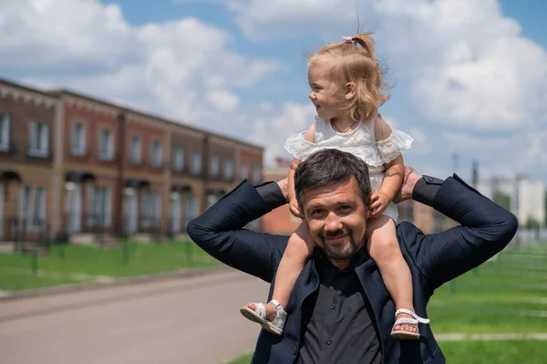 Усміхнений чоловік у костюмі гуляє зі своєю дочкою на відкритому повітрі. Маленька дівчинка в білій сукні сидить на плечах турботливого тата. Щасливе батьківство . — стокове фото