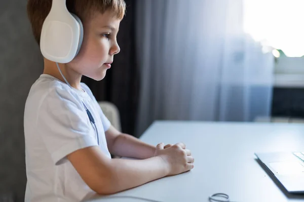 Un niño diligente en auriculares se sienta en un escritorio y mira una lección en línea en un ordenador portátil. — Foto de Stock