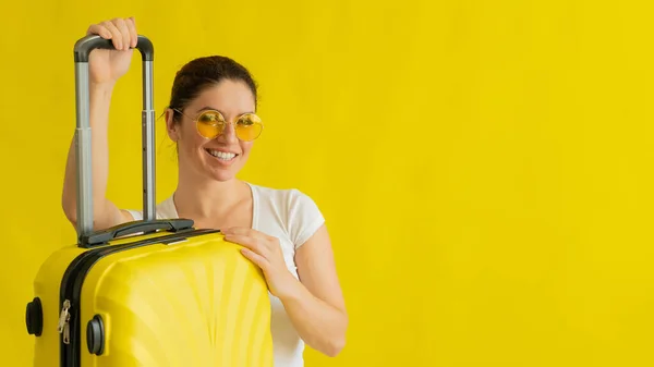戴着太阳镜，拿着黄色背景可伸缩手柄的手提箱微笑的女人 — 图库照片