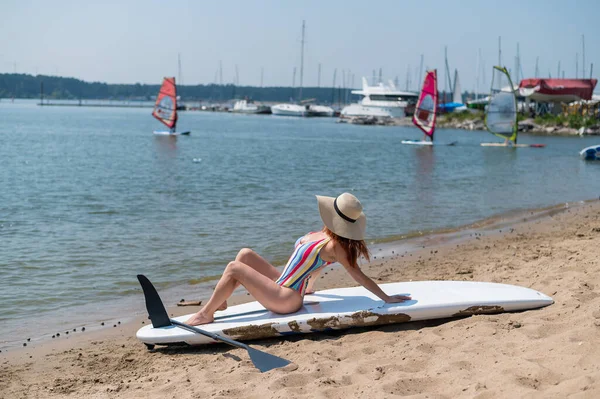 Una mujer en traje de baño y un sombrero posan en una tabla de sup en la playa. — Foto de Stock