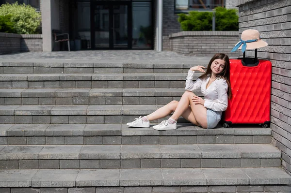 Eine schöne kaukasische Frau mit Hut und kurzen Hosen sitzt mit einem großen roten Koffer auf der Treppe. Lächelndes Mädchen, das auf ein Taxi wartet. Reisekonzept. — Stockfoto