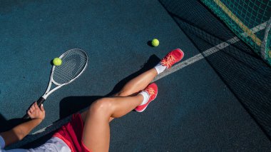 Spor etekli yüzü olmayan bir kız tenis kortunda oturuyor ve elinde bir roket tutuyor. Kadın bacaklarının üst görüntüsü.
