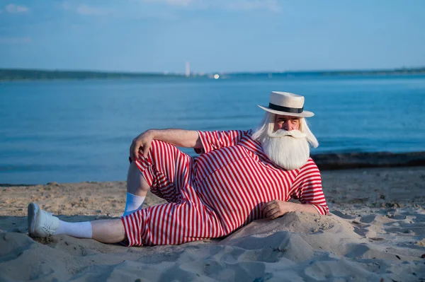 Un uomo anziano in costume da bagno retrò a righe prende il sole sulla spiaggia. Un vecchio uomo barbuto dai capelli grigi con un cappello giace sulla sabbia vicino al mare. — Foto Stock