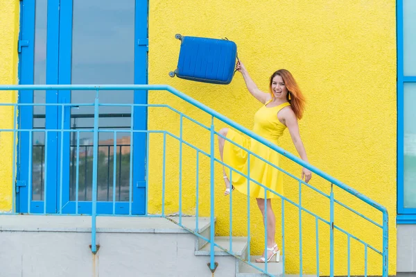 幸せな赤髪の女性でA黄色のドレス行きます2階にブルースーツケースを保持. — ストック写真