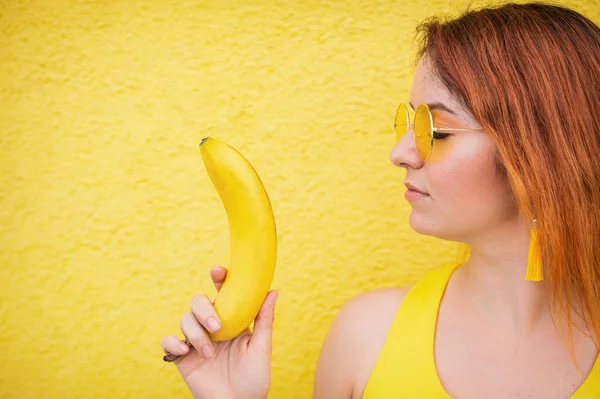 Porträtt av den kaukasiska rödhåriga kvinnan i solglasögon med en banan i händerna. Vacker förförisk flicka poserar på en gul bakgrund. — Stockfoto