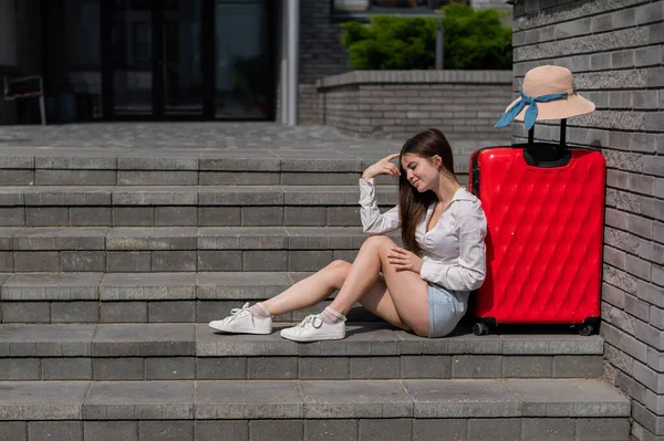 Eine schöne kaukasische Frau mit Hut und kurzen Hosen sitzt mit einem großen roten Koffer auf der Treppe. Lächelndes Mädchen, das auf ein Taxi wartet. Reisekonzept. — Stockfoto