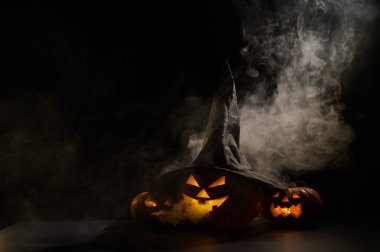 Sisin ortasında karanlıkta üç tane balkabağı feneri parlıyor. Cadılar Bayramı kabağı ve cadı şapkası..