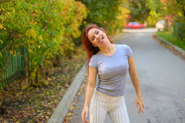 Uma bela mulher ruiva está andando pela rua e dançando. Menina feliz em uma camiseta cinza e leggings listrados caminha e se diverte no parque de outono. Foto em movimento. — Fotografia de Stock