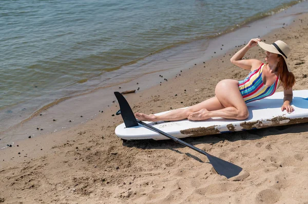 Eine Frau in Badeanzug und Hut posiert auf einem Surfbrett am Strand. — Stockfoto