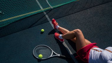 Spor etekli yüzü olmayan bir kız tenis kortunda oturuyor ve elinde bir roket tutuyor. Kadın bacaklarının üst görüntüsü.