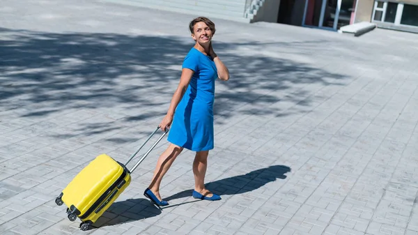 Mujer feliz caminando por la calle sosteniendo maleta amarilla con ruedas — Foto de Stock