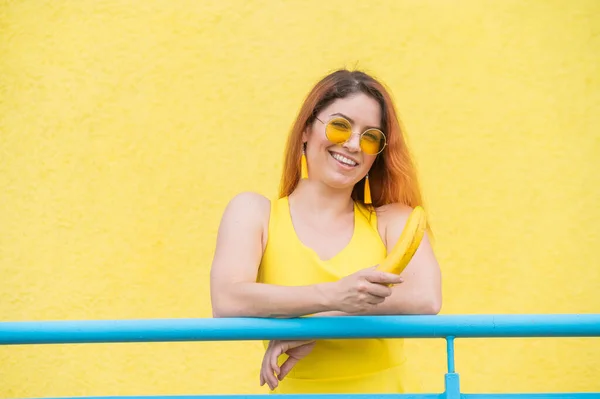 Červenovlasá žena v slunečních brýlích drží banán na žlutém pozadí. — Stock fotografie