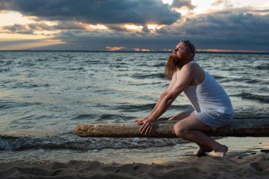 Gün batımında sahilde poz veren kızıl sakallı komik kel adam. Çekici bir kızın esprili erkek parodisi..