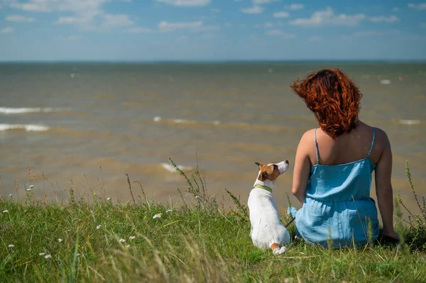 Kobieta siedzi na brzegu stawu z psem. Widok z tyłu dziewczyny na wakacjach w przyrodzie ze zwierzątkiem domowym. — Zdjęcie stockowe