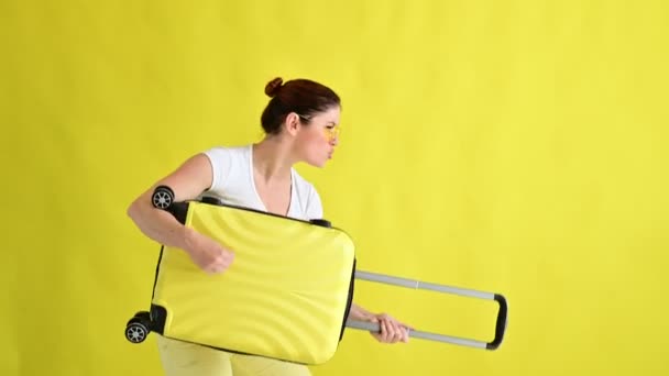 Glimlachende vrouw met een zonnebril die rondhangt en een koffer vasthoudt als een gitaar op een gele achtergrond. Een opgewonden meisje in afwachting van een zomervakantie simuleert het bespelen van een snaarinstrument. — Stockvideo