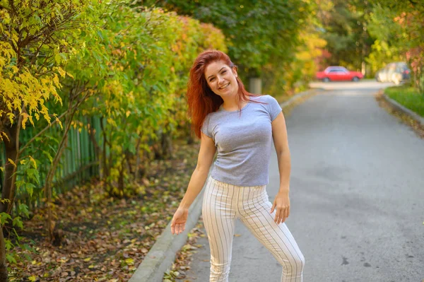 En vacker rödhårig kvinna går längs gatan och dansar. Glad tjej i grå T-shirt och randiga leggings promenader och har kul i höstparken. Foto i rörelse. — Stockfoto