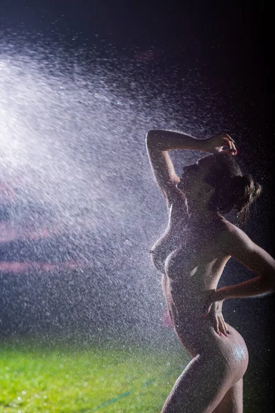 Uma bela mulher nua europeia fica em um gramado sob aspersão à noite. Flashes iluminar uma menina sem roupas em gotas de respingos de grama de água. — Fotografia de Stock