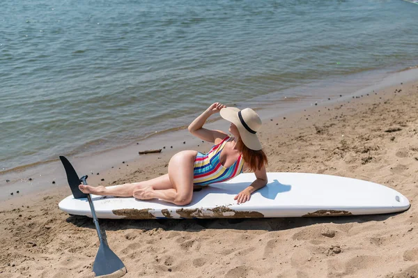 Eine Frau in Badeanzug und Hut posiert auf einem Surfbrett am Strand. — Stockfoto