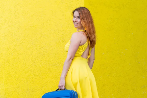 Женщина в жёлтом платье разворачивается и держит синий чемодан на цветном фоне — стоковое фото
