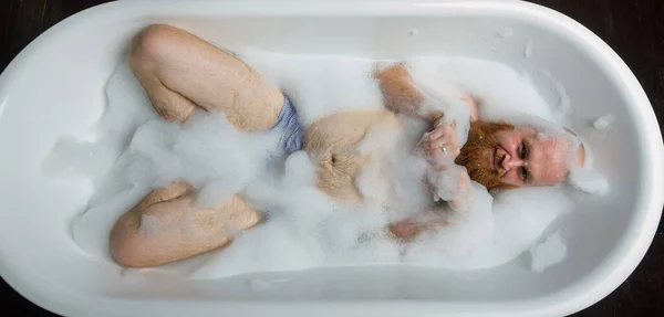 Vista dall'alto di un uomo calvo con una barba rossa che schizza nel bagno di schiuma. Una foto divertente. Una parodia di ragazze glamour. — Foto Stock