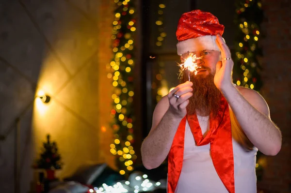 Santa con destellos navideños. Un hombre con una barba roja en un sombrero de Santa Claus y una camiseta para un bengalas de quemaduras alcohólicas. Parodia. —  Fotos de Stock