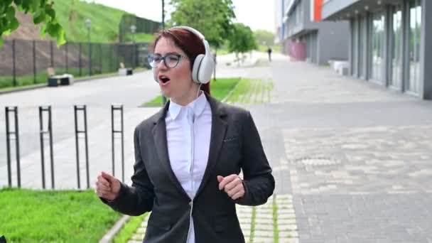 Blanke zakenvrouw in een pak luisterend naar muziek met koptelefoon en meezingend om buiten te dansen — Stockvideo