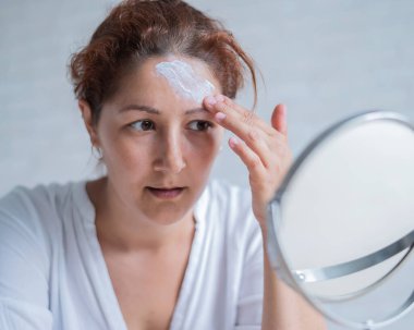 Vitiligo 'su olan beyaz bir kadının portresi güneş kremi kullanıyor. Alnında beyaz pigment lekesi olan bir kız aynaya bakar ve krem sürmüştür..