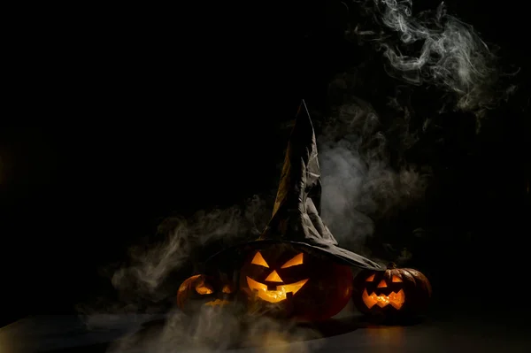 Three jack o lanterns glow in the dark amidst the fog. Halloween pumpkin in a witch hat.