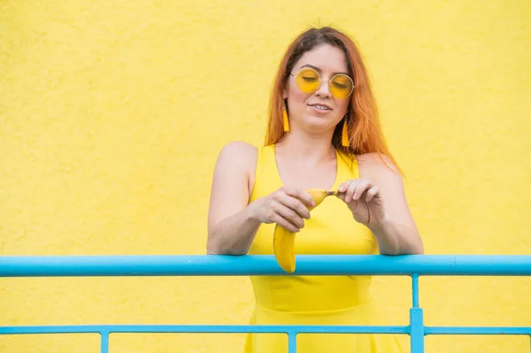 Рыжая женщина в солнечных очках держит банан на желтом фоне. — стоковое фото