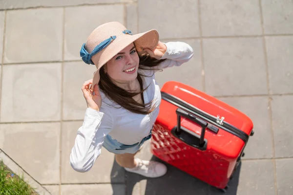 穿着夏帽、头戴红色手提箱的年轻漂亮的高加索女人的头像。快乐的微笑的女孩带着一个大旅行袋去度假。在炎热的晴天穿短裤的迷人女士. — 图库照片