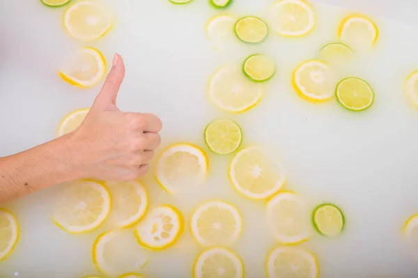 A womans hand shows her thumb against the background of a milk bath with citrus fruits. Woman gestures approval while taking a bath with lemons and limes. — Stock Photo, Image