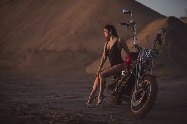 Hermosa mujer sexy en traje de encaje y tacones altos se sienta en una motocicleta en un fondo industrial — Foto de Stock