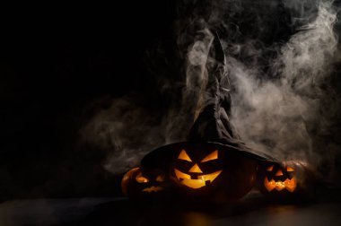 Sisin ortasında karanlıkta üç tane balkabağı feneri parlıyor. Cadılar Bayramı kabağı ve cadı şapkası..